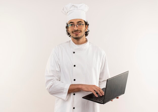 Cocinero de sexo masculino joven sonriente que lleva el uniforme del cocinero y las gafas que sostienen el ordenador portátil aislado en la pared blanca
