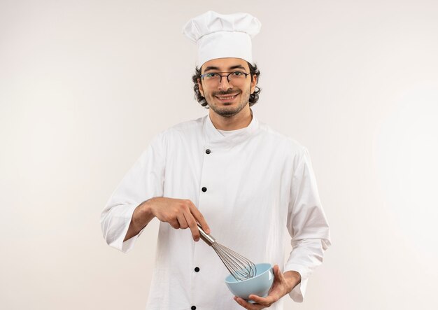 Cocinero de sexo masculino joven sonriente que lleva el uniforme del cocinero y los vidrios que sostienen el batidor y el cuenco aislado en la pared blanca