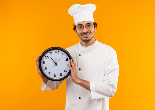 Cocinero de sexo masculino joven sonriente que lleva el uniforme del cocinero y los vidrios que sostienen el reloj de pared aislado en la pared amarilla