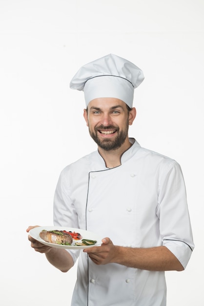 Cocinero de sexo masculino joven sonriente en el uniforme del cocinero que sostiene el plato preparado de la carne de vaca contra el fondo blanco