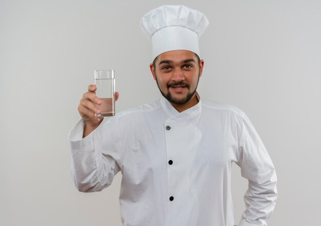 Cocinero de sexo masculino joven sonriente en uniforme del cocinero que sostiene el vaso de agua aislado en el espacio en blanco