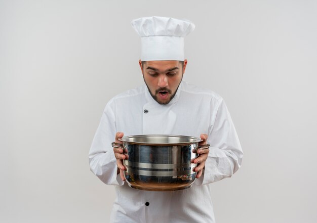 Cocinero de sexo masculino joven sorprendido en uniforme del cocinero que sostiene y que mira dentro de la olla aislada en el espacio en blanco