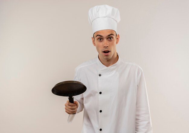 Cocinero de sexo masculino joven sorprendido vistiendo uniforme del cocinero que sostiene la sartén con el espacio de la copia