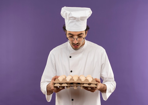 Cocinero de sexo masculino joven sorprendido vistiendo uniforme del cocinero y los vidrios que sostienen el lote de huevos en púrpura