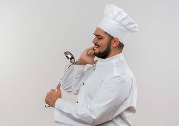 Cocinero de sexo masculino joven en uniforme de chef sosteniendo la cuchara y usándolo como espejo aislado en el espacio rosa