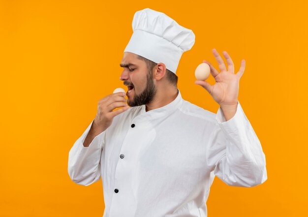 Cocinero de sexo masculino joven en uniforme de chef sosteniendo y tratando de morder el huevo aislado en el espacio naranja