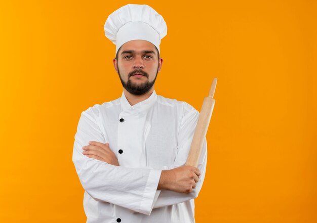 Cocinero de sexo masculino joven en uniforme del cocinero que se coloca con la postura cerrada y que sostiene el rodillo