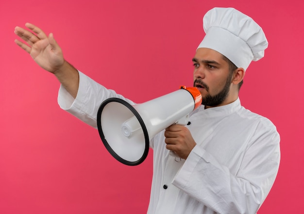 Cocinero de sexo masculino joven en uniforme del cocinero que habla por el altavoz con la mano levantada y mirando al lado