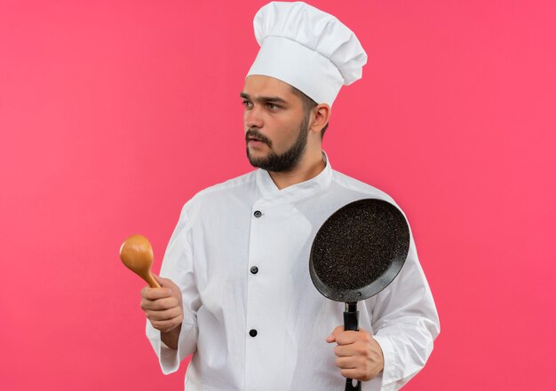 Cocinero de sexo masculino joven en uniforme del cocinero que sostiene la cuchara y la sartén que mira el lado aislado en el espacio rosado