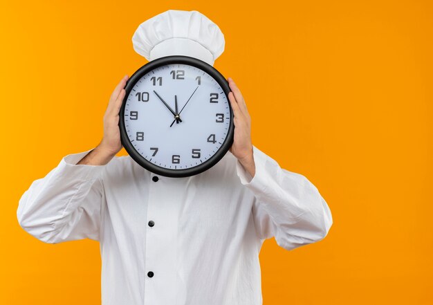 Cocinero de sexo masculino joven en uniforme del cocinero que sostiene y que se esconde detrás del reloj aislado en el espacio anaranjado