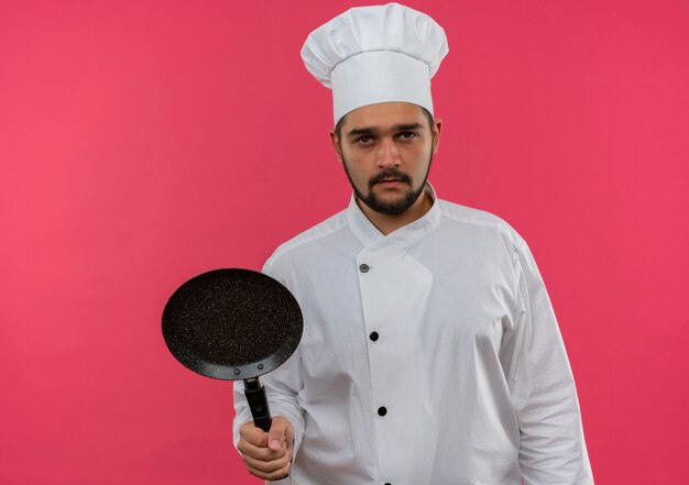 Cocinero de sexo masculino joven en uniforme del cocinero que sostiene la sartén mirando aislado en el espacio rosado