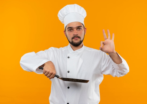 Cocinero de sexo masculino joven en uniforme del cocinero que sostiene la sartén y que hace el signo bien aislado en el espacio naranja