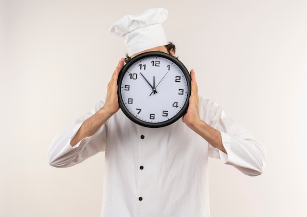 Cocinero de sexo masculino joven vistiendo uniforme de chef y anteojos cubiertos con reloj de pared aislado en la pared blanca