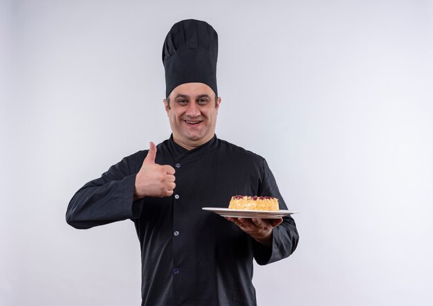 Cocinero de sexo masculino de mediana edad complacido en uniforme de chef sosteniendo la torta en la placa con el pulgar hacia arriba en la pared blanca aislada con espacio de copia