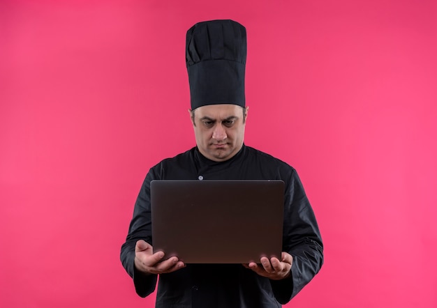 Cocinero de sexo masculino de mediana edad confundido en uniforme del cocinero que mira la computadora portátil en su mano en la pared rosada aislada con el espacio de la copia