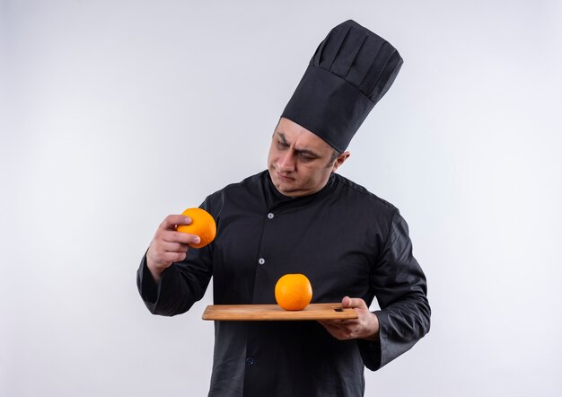 Cocinero de sexo masculino de mediana edad confundido en uniforme del cocinero que sostiene la naranja en la tabla de cortar y que mira la naranja en su mano en la pared blanca aislada