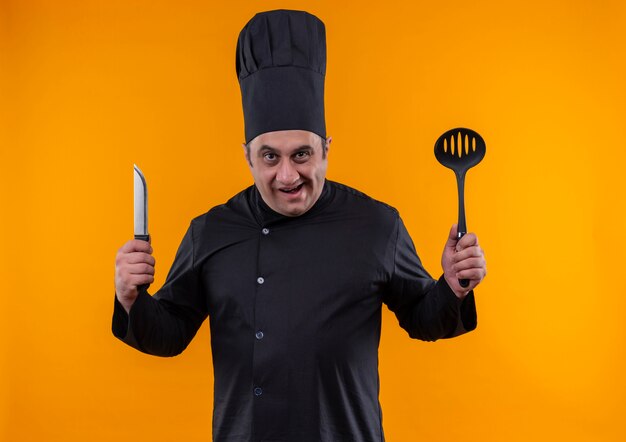 Cocinero de sexo masculino de mediana edad enojado en uniforme del cocinero que sostiene la cuchara y el cuchillo en la pared amarilla