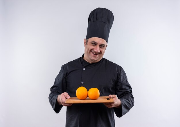 Cocinero de sexo masculino de mediana edad sonriente en uniforme del cocinero que sostiene las naranjas en la tabla de cortar