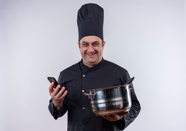 Cocinero de sexo masculino de mediana edad sonriente en uniforme del cocinero que sostiene la olla y el teléfono