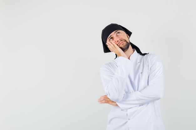 Cocinero de sexo masculino que se inclina la mejilla en la palma levantada en uniforme blanco y que parece pensativo. vista frontal.