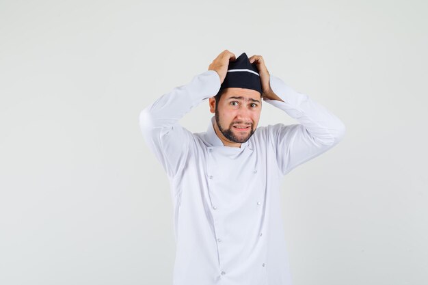 Cocinero de sexo masculino que lleva a cabo la mano en la cabeza en uniforme blanco y que parece preocupado. vista frontal.