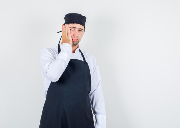 Cocinero de sexo masculino que lleva a cabo la mano en la mejilla en uniforme, delantal y mirando desilusionado, vista frontal