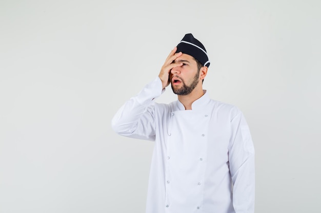 Cocinero de sexo masculino que lleva a cabo la mano en su cara en uniforme blanco y que parece preocupado, vista frontal.