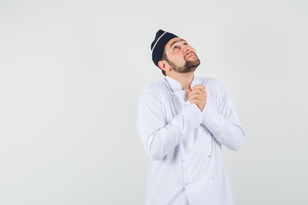 Cocinero de sexo masculino que lleva a cabo las manos juntas en gesto de oración en uniforme blanco y mirando esperanzado, vista frontal.