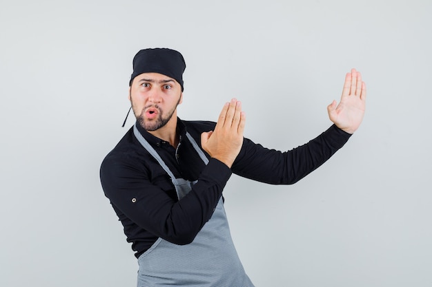 Cocinero de sexo masculino que muestra el gesto de chuleta de karate en camisa, delantal y con enojo. vista frontal.