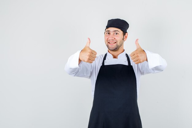 Cocinero de sexo masculino que muestra los pulgares para arriba en uniforme, delantal y que parece feliz. vista frontal.