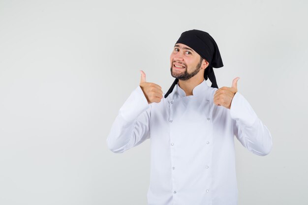 Cocinero de sexo masculino que muestra los pulgares dobles para arriba en uniforme blanco y que mira positiva, vista frontal.