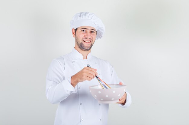 Cocinero de sexo masculino que sostiene el batidor y el cuenco en uniforme blanco y que parece alegre.