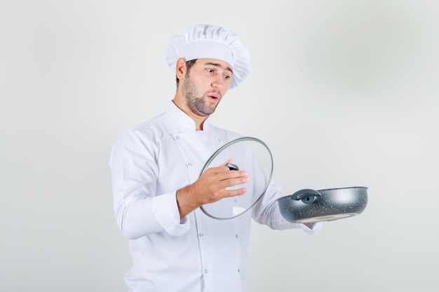 Cocinero de sexo masculino que sostiene la cacerola y la tapa de cristal en uniforme blanco y que parece sorprendido.