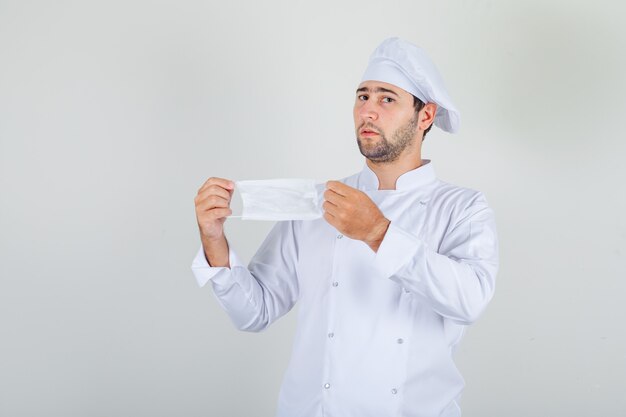 Cocinero de sexo masculino que sostiene la máscara médica en uniforme blanco y parece vacilante.