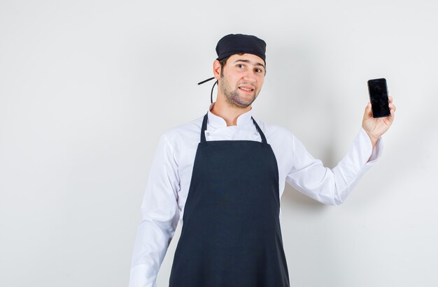 Cocinero de sexo masculino que sostiene el teléfono móvil en uniforme, delantal y que parece optimista. vista frontal.