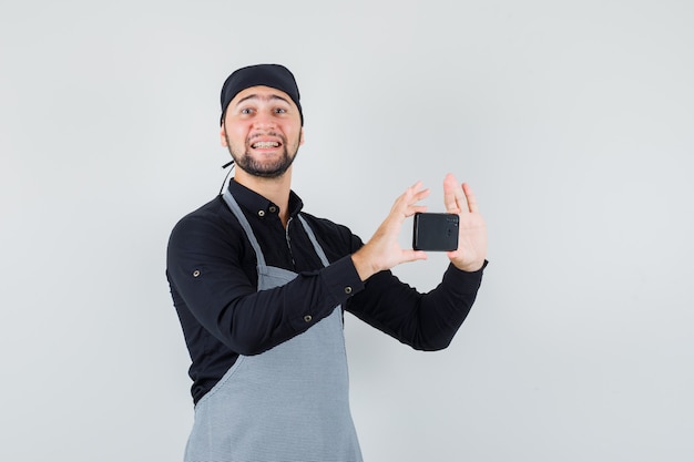 Cocinero de sexo masculino que toma la foto en el teléfono móvil en camisa, delantal y mirando alegre. vista frontal.