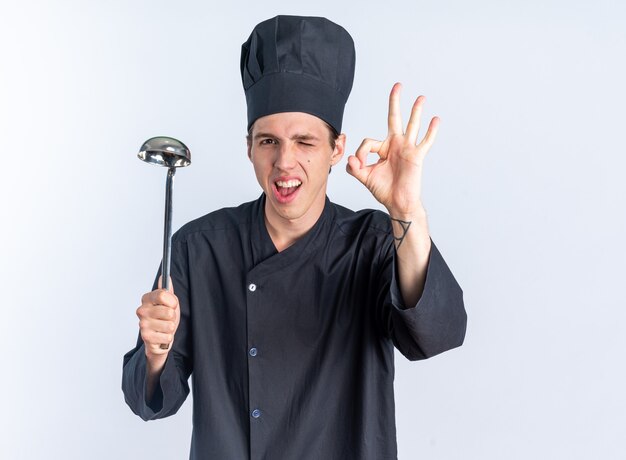 Cocinero de sexo masculino rubio joven alegre en uniforme del cocinero y la tapa que sostiene el cucharón que hace el guiño de signo ok