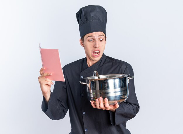 Cocinero de sexo masculino rubio joven asustado en uniforme del cocinero y la tapa que sostiene el bloc de notas y la olla mirando dentro de la olla aislada en la pared blanca