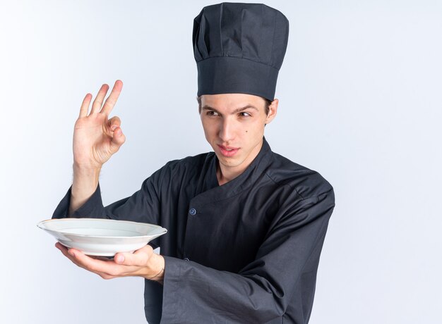 Cocinero de sexo masculino rubio joven confiado en el uniforme del cocinero y la tapa que mira el lado que se extiende la placa que hace la muestra aceptable aislada en la pared blanca