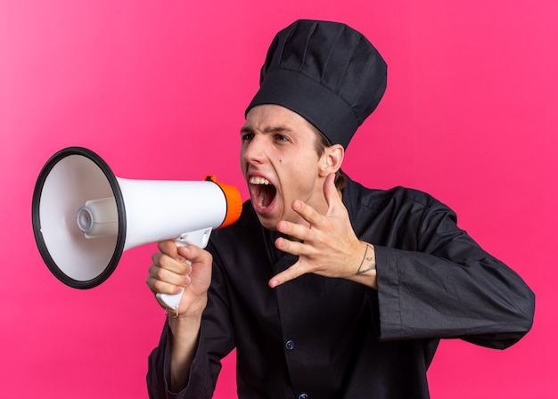 Cocinero de sexo masculino rubio joven furioso en uniforme del cocinero y gorra mirando al lado gritando en altavoz manteniendo la mano en el aire aislado en la pared rosa