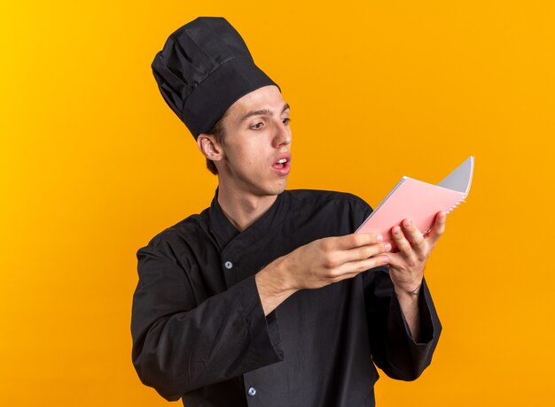 Cocinero de sexo masculino rubio joven impresionado en el bloc de notas de la lectura de la tapa y del uniforme del cocinero aislado en la pared naranja