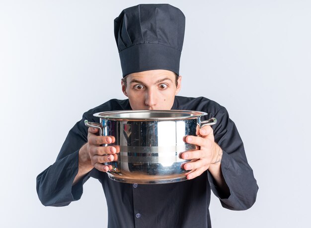 Cocinero de sexo masculino rubio joven sorprendido en uniforme del cocinero y la tapa que sostiene y que mira la olla