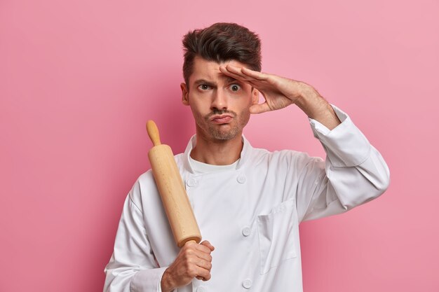 Cocinero de sexo masculino serio sostiene el rodillo, mantiene la mano en la frente, piensa qué cocinar para los visitantes del restaurante