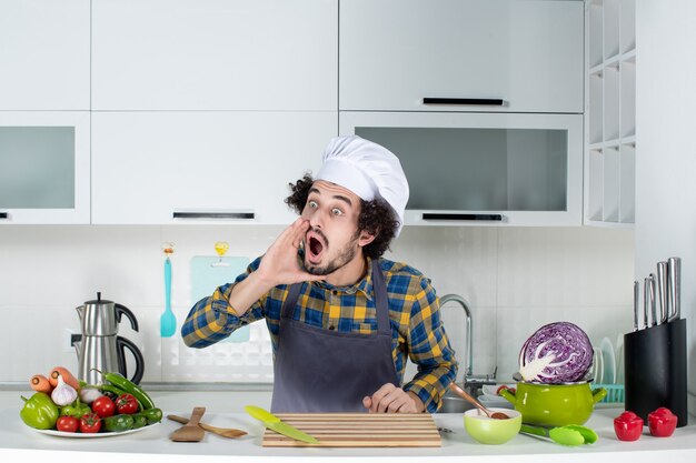 Cocinero de sexo masculino sorprendido con verduras frescas y cocinando con utensilios de cocina y llamando a alguien en la cocina blanca