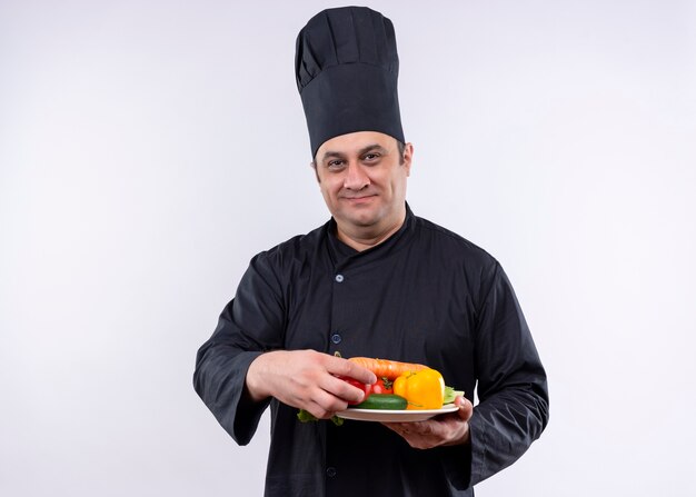 Cocinero de sexo masculino con uniforme negro y sombrero de cocinero sosteniendo un plato con verduras frescas mirando a la cámara con una sonrisa en la cara de pie sobre fondo blanco