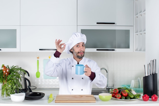 Cocinero de sexo masculino de la vista frontal que sostiene el sombrero del cocinero que se coloca detrás de la mesa de la cocina