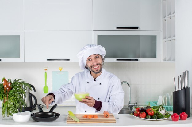 Cocinero de sexo masculino de la vista frontal en uniforme que sostiene el cuenco apuntando a las cacerolas en la cocina