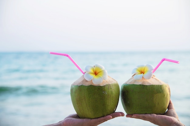 Coco fresco en manos de los pares con plumeria adornado en la playa con la onda del mar - turista de los pares de la luna de miel con concepto de las vacaciones del sol de la fruta fresca y de la arena de mar