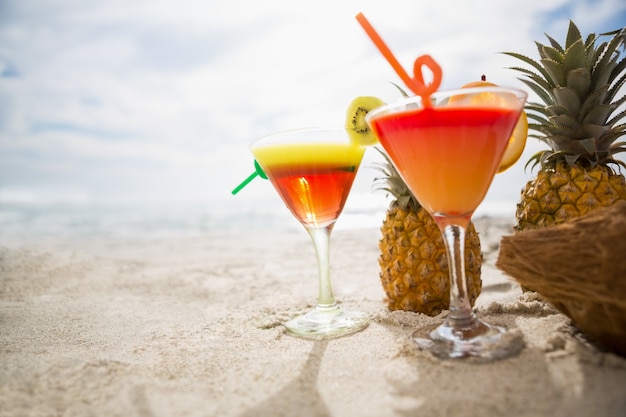Coco, piña y dos vasos de cóctel de bebidas mantienen en la arena en la playa tropical