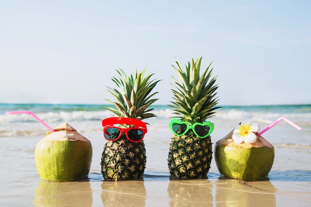 El coco y la piña frescos ponen los vidrios encantadores del sol en la playa limpia de la arena con la onda del mar - fruta fresca con concepto de las vacaciones del sol de la arena de mar